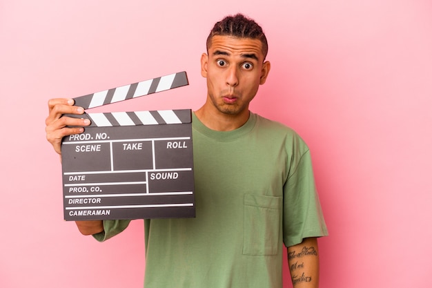 Young venezuelan man holding a clapperboard isolated on pink background shrugs shoulders and open eyes confused.