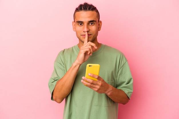 Young Venezuelan man holding a mobile phone isolated on pink wall keeping a secret or asking for silence.