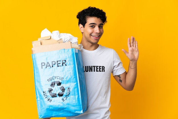 Young Venezuelan man holding a recycling bag full of paper to recycle saluting with hand with happy expression