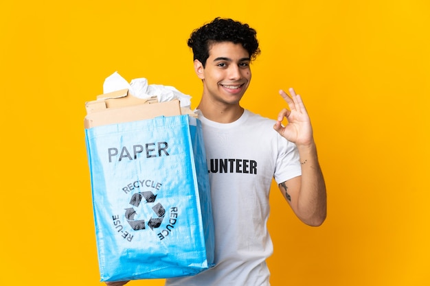 Молодой венесуэльский мужчина держит мешок для переработки, полный бумаги для переработки, показывает пальцами знак ОК