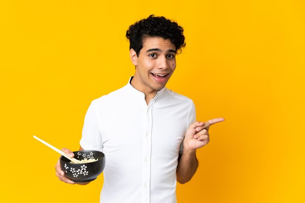 Young Venezuelan man on yellow surprised and pointing finger to the side while holding a bowl of noodles with chopsticks