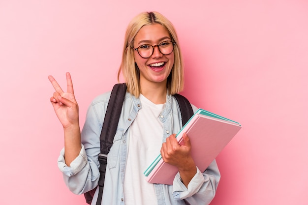 ピンクの壁に孤立した若いベネズエラの学生女性は、指で平和のシンボルを示して楽しくてのんきです。