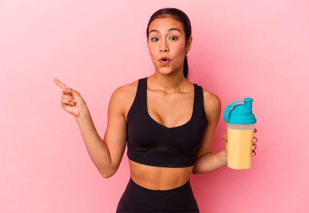 Young Venezuelan woman drinking a protein shake isolated on pink background pointing to the side