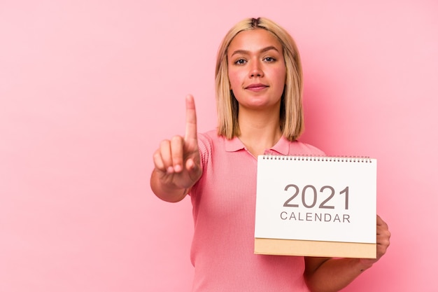 Giovane donna venezuelana che tiene un calendario isolato sulla parete rosa che mostra il numero uno con il dito