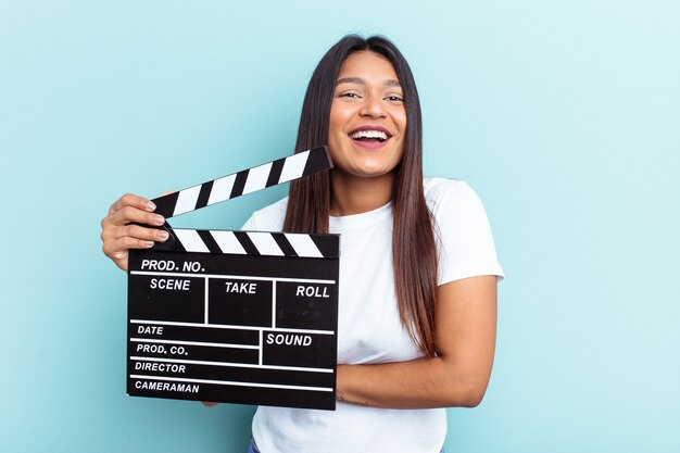 Young Venezuelan woman holding a clapperboard isolated on blue background laughing and having fun.