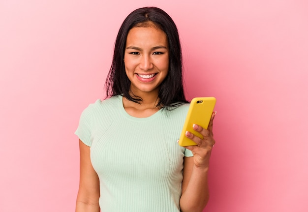 ピンクの背景に分離された携帯電話を持っている若いベネズエラの女性は幸せ、笑顔、陽気な。