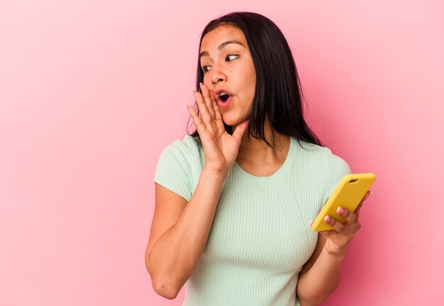 Young Venezuelan woman holding a mobile phone isolated on pink background is saying a secret hot braking news and looking aside