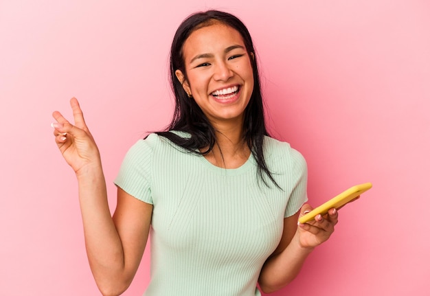 ピンクの背景に分離された携帯電話を持っている若いベネズエラの女性は、指で平和のシンボルを示す喜びと屈託のない