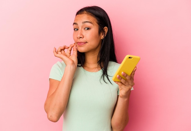 Young venezuelan woman holding a mobile phone isolated on pink\
background with fingers on lips keeping a secret