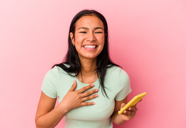 Young Venezuelan woman holding a mobile phone isolated on pink wall laughs out loudly keeping hand on chest.