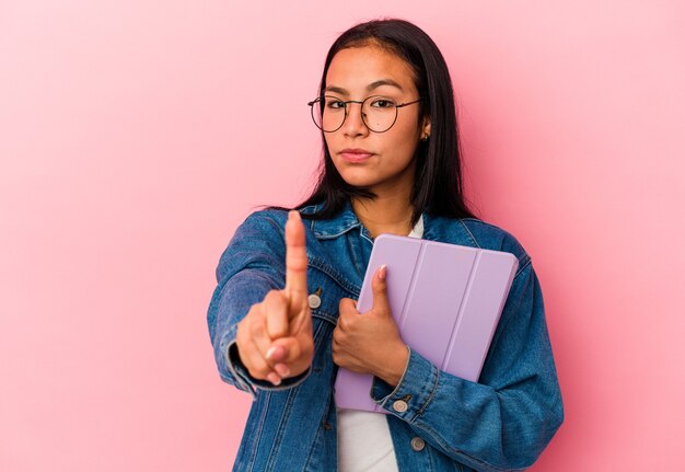 ピンクの背景に分離されたタブレットを持っている若いベネズエラの女性は、指でナンバーワンを示しています。