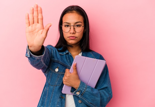 ピンクの背景に分離されたタブレットを保持している若いベネズエラの女性は、一時停止の標識を示している手を伸ばして立って、あなたを防ぎます。