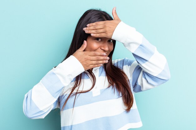 Young Venezuelan woman isolated on blue background blink at the camera through fingers, embarrassed covering face.