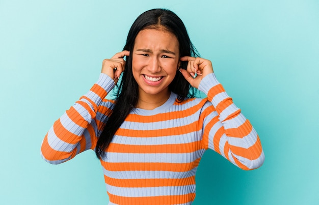 Young Venezuelan woman isolated on blue background covering ears with fingers, stressed and desperate by a loudly ambient.