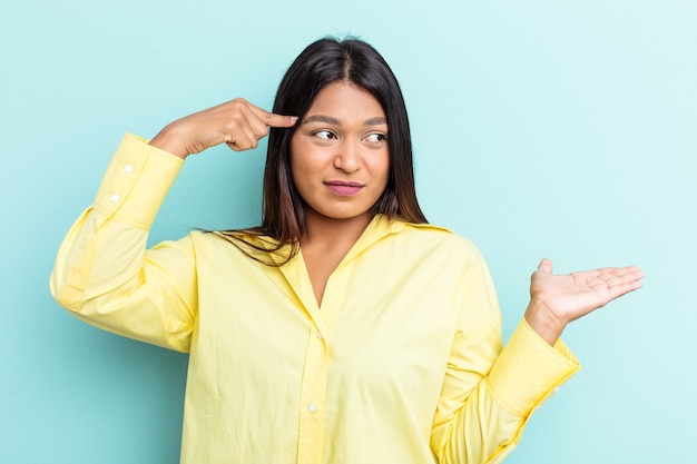 Young Venezuelan woman isolated on blue background holding and showing a product on hand.