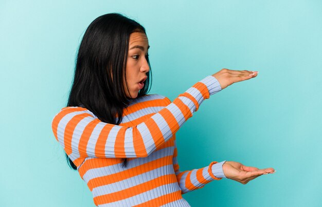 Young Venezuelan woman isolated on blue background shocked and amazed holding a copy space between hands.