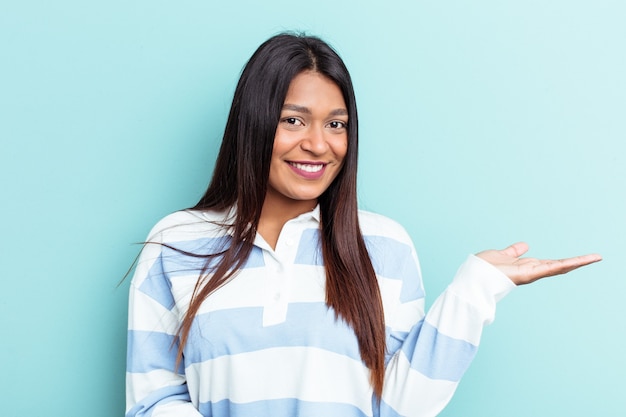 Young Venezuelan woman isolated on blue background showing a copy space on a palm and holding another hand on waist.