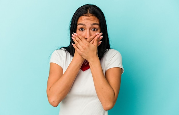 Young Venezuelan woman isolated on blue wall covering mouth with hands looking worried.