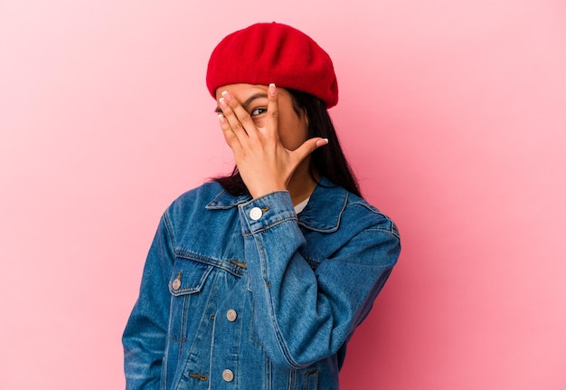 Young Venezuelan woman isolated on pink background blink at the camera through fingers embarrassed covering face