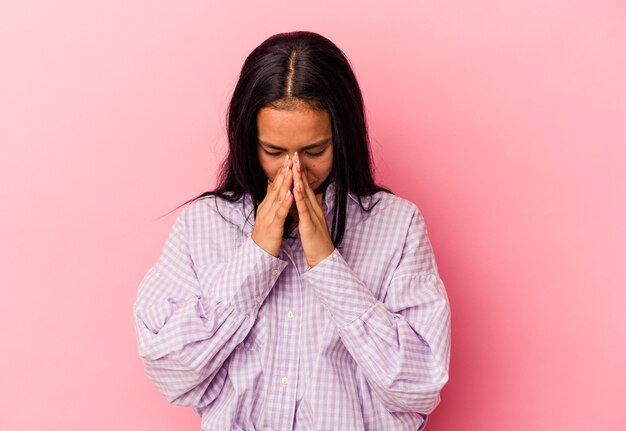 Young Venezuelan woman isolated on pink background praying, showing devotion, religious person looking for divine inspiration.