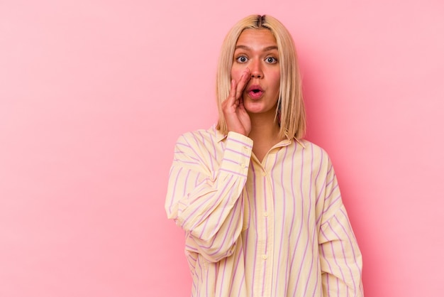 Young venezuelan woman isolated on pink wall is saying a secret hot braking news and looking aside