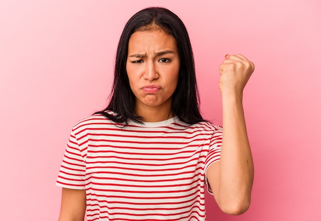 Young Venezuelan woman isolated on pink wall showing fist, aggressive facial expression.