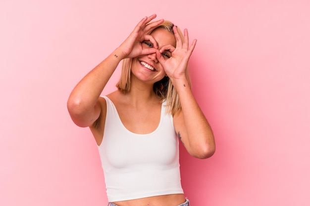 Young venezuelan woman isolated on pink wall showing okay sign over eyes