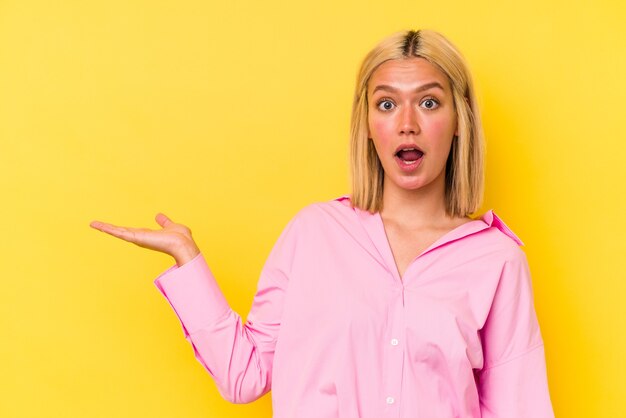 Young venezuelan woman isolated on yellow background impressed holding copy space on palm