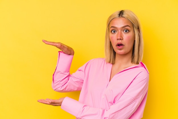 Young venezuelan woman isolated on yellow background shocked and amazed holding a copy space between hands.