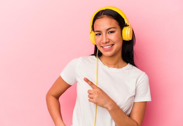 ピンクの背景に分離された音楽を聞いている若いベネズエラの女性は、笑顔で脇を指して、空白のスペースで何かを示しています。