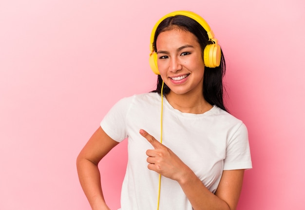 Young Venezuelan woman listening to music isolated on pink background smiling and pointing aside, showing something at blank space.