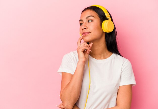 Young Venezuelan woman listening to music isolated on pink wall relaxed thinking about something looking at a copy space.