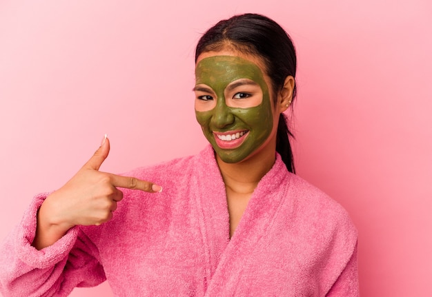 Young Venezuelan woman wearing a bathrobe and facial mask isolated on pink background person pointing by hand to a shirt copy space, proud and confident