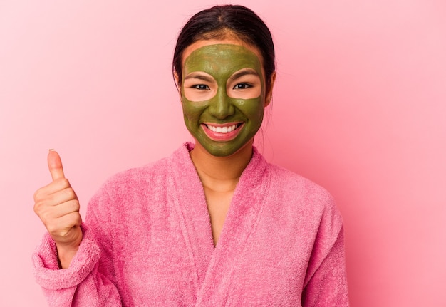 Photo young venezuelan woman wearing a bathrobe and facial mask isolated on pink background smiling and raising thumb up