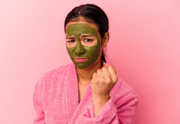 Young Venezuelan woman wearing a bathrobe and facial mask isolated on pink wall showing fist to camera, aggressive facial expression.