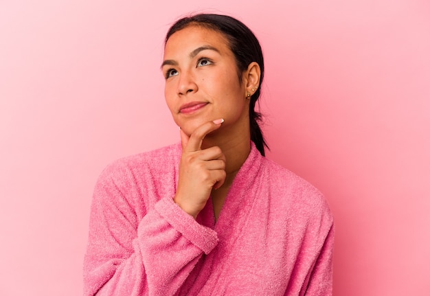 Young Venezuelan woman wearing a bathrobe isolated on pink wall looking sideways with doubtful and skeptical expression.