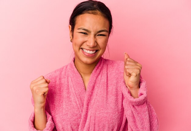 Young Venezuelan woman wearing a bathrobe isolated on pink wall raising fist after a victory, winner concept.