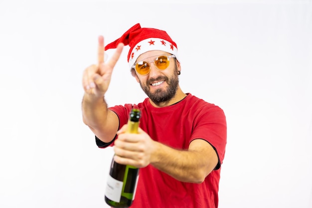 Young very happy Caucasian man with red Christmas hat toasting with a bottle of champagne on a white background, dressed in a red t-shirt and sunglasses