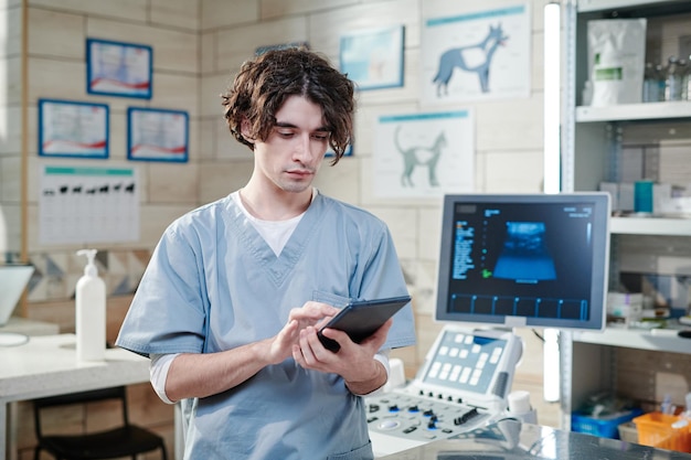 Young vet doctor in uniform typing on digital tablet and working online while standing at his vet office