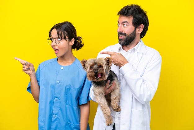 Young veterinarian couple with dog isolated on yellow background surprised and pointing finger to the side