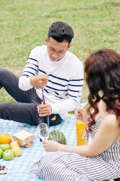 Young Vietnamese man opening bottle of wine for his girlfriend when they are enjoying romantic picnic