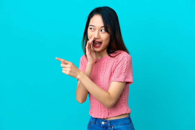 Young vietnamese woman isolated on blue background pointing to the side to present a product and whispering something