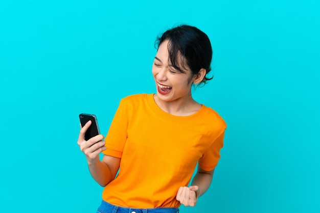 Young Vietnamese woman isolated on blue background using mobile phone and doing victory gesture