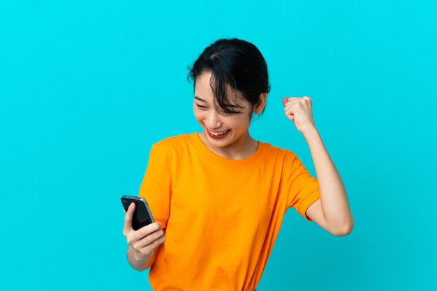 Young Vietnamese woman isolated on blue background using mobile phone and doing victory gesture