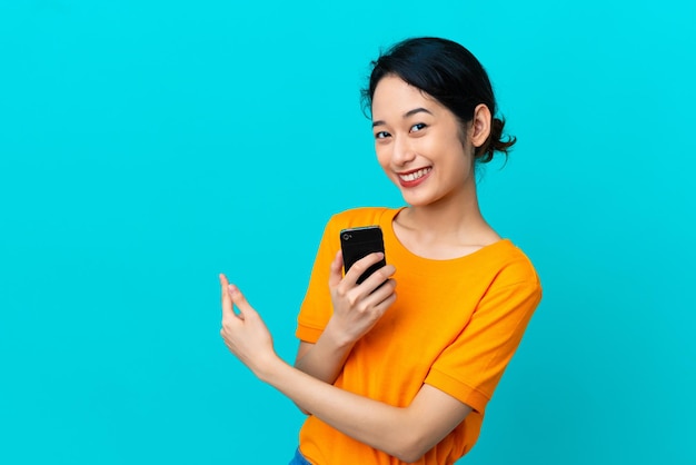 Young Vietnamese woman isolated on blue background using mobile phone and pointing back