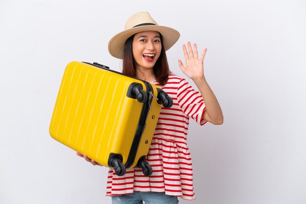 Giovane donna vietnamita isolata su sfondo bianco in vacanza con valigia da viaggio e saluto