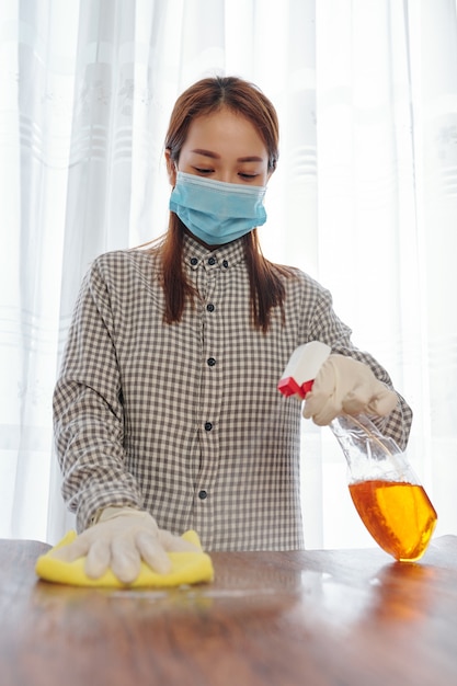 Young Vietnamese woman in medical mask using cleaning detergent when wiping furniture in her apartment