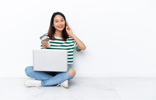 Young Vietnamese woman with a laptop sitting on the floor isolated on white wall holding coffee to take away and a mobile