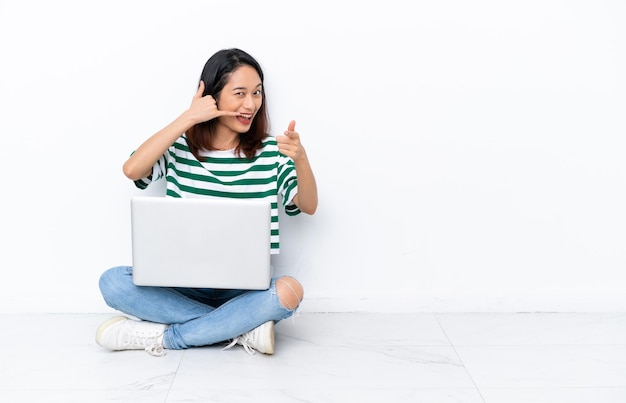 Young Vietnamese woman with a laptop sitting on the floor isolated on white wall making phone gesture and pointing front