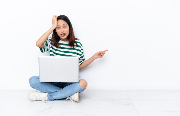 Young Vietnamese woman with a laptop sitting on the floor isolated on white wall surprised and pointing finger to the side